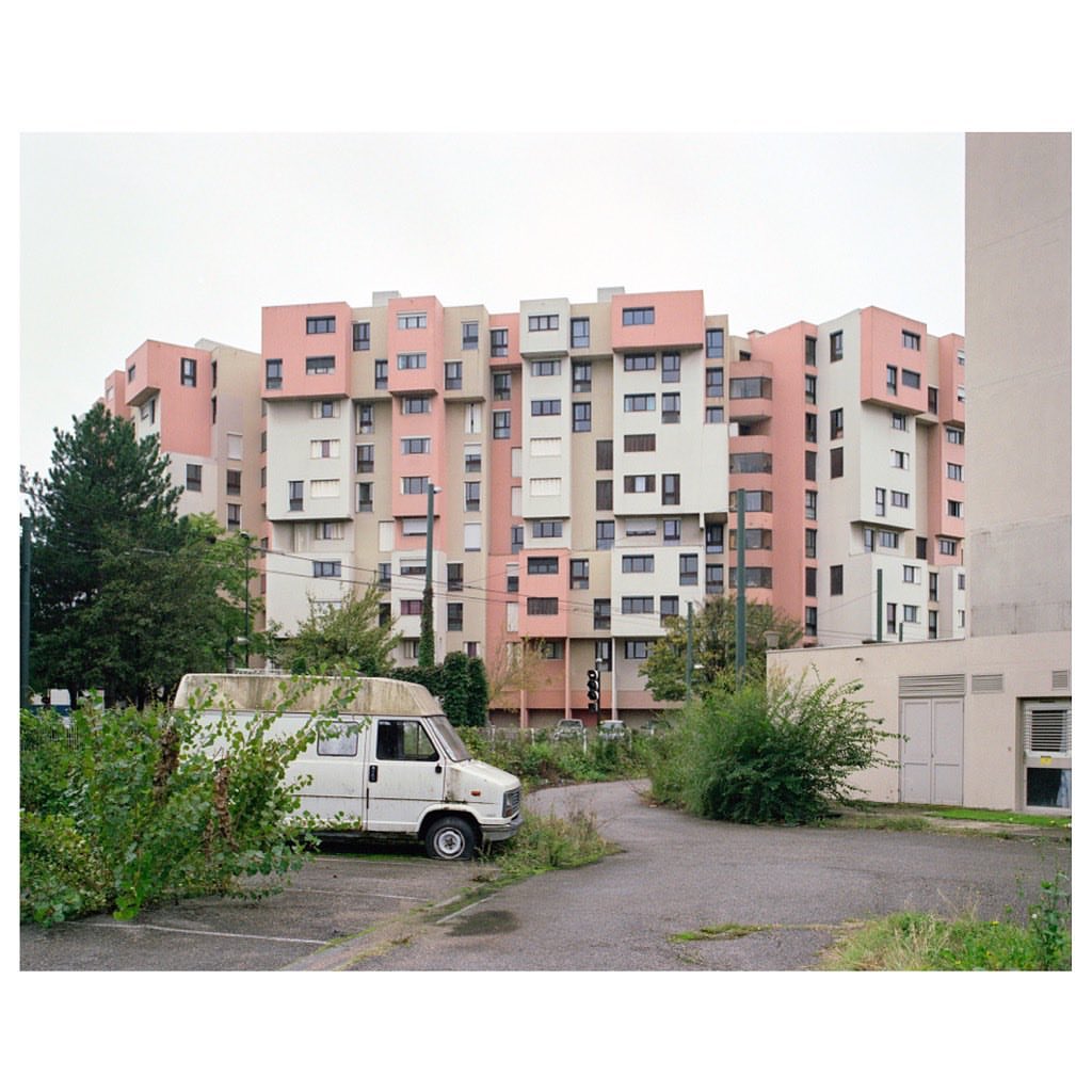 Residential apartment building featuring pink and beige exterior, parked van, and green vegetation in an urban environment.