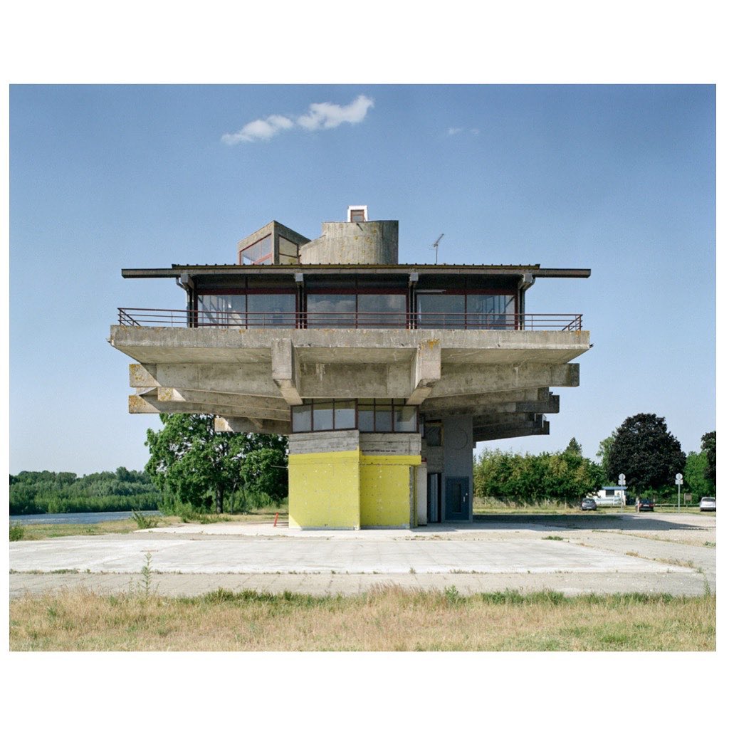 Architectural photo of a concrete tower with a yellow base and a flat rooftop terrace against a backdrop of trees and a blue sky.