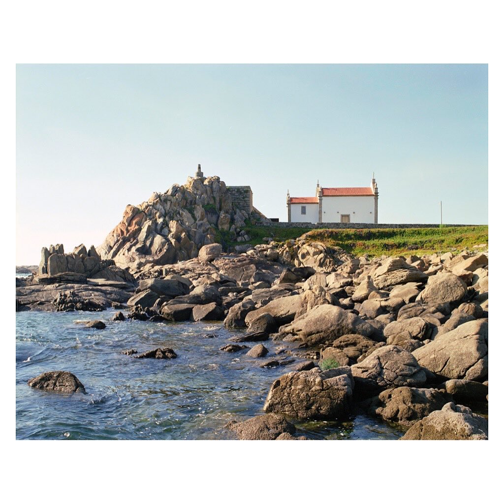 Coastal landscape featuring rocky shoreline, blue water, and a quaint white chapel with red roof tiles on a grassy hill under clear skies.