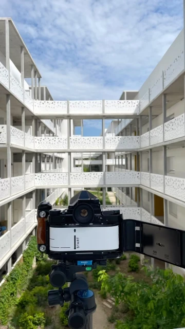 Analog camera mounted on a tripod faces modern architecture with a courtyard, showcasing a blend of technology and contemporary design.