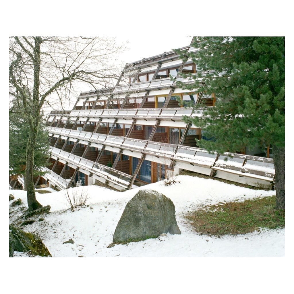 Exterior view of an A-frame chalet with a unique staggered design, set in a snowy landscape with trees and a large rock in the foreground.