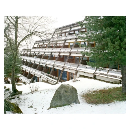 Exterior view of an A-frame chalet with a unique staggered design, set in a snowy landscape with trees and a large rock in the foreground.