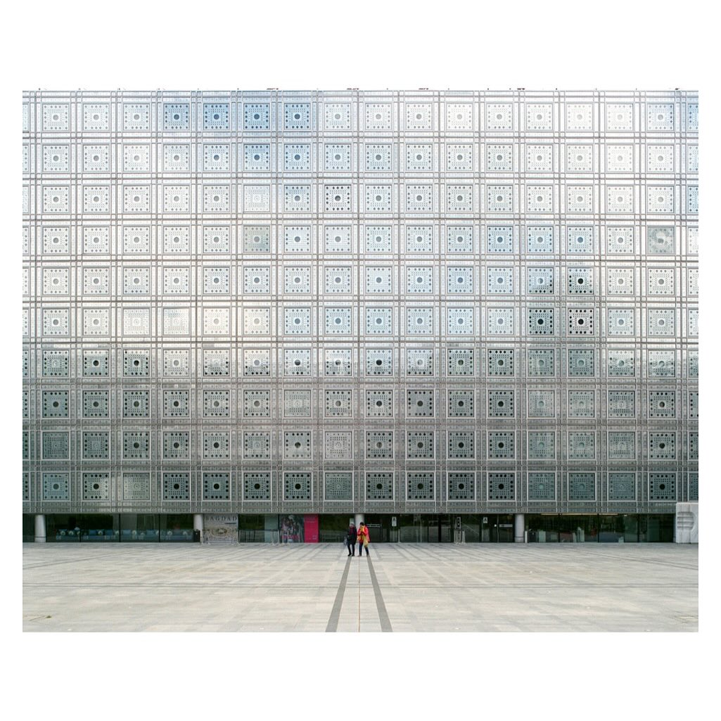 Modern architecture featuring an ornate patterned facade and a large, open plaza with people walking in the urban environment.