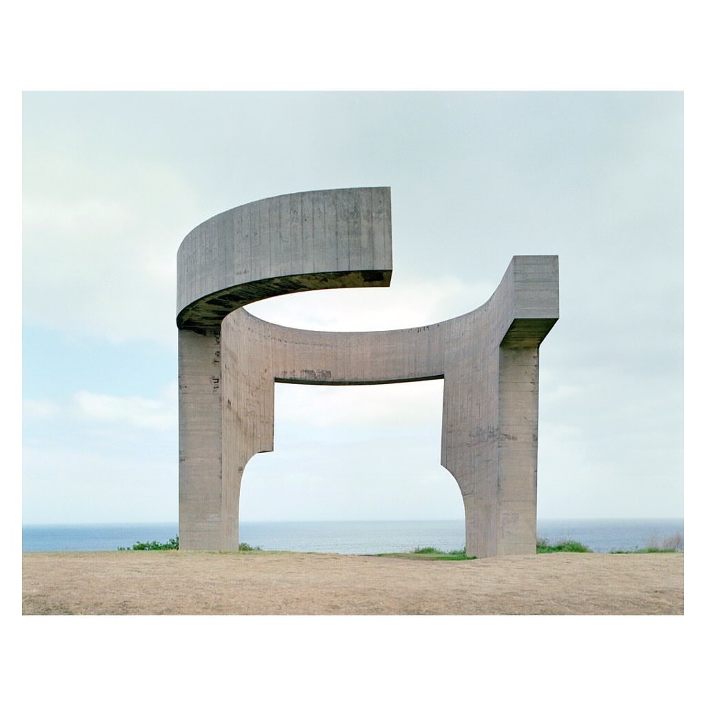 Modern outdoor concrete architectural sculpture stands on a hilltop overlooking the ocean in overcast daylight, creating visual interest.