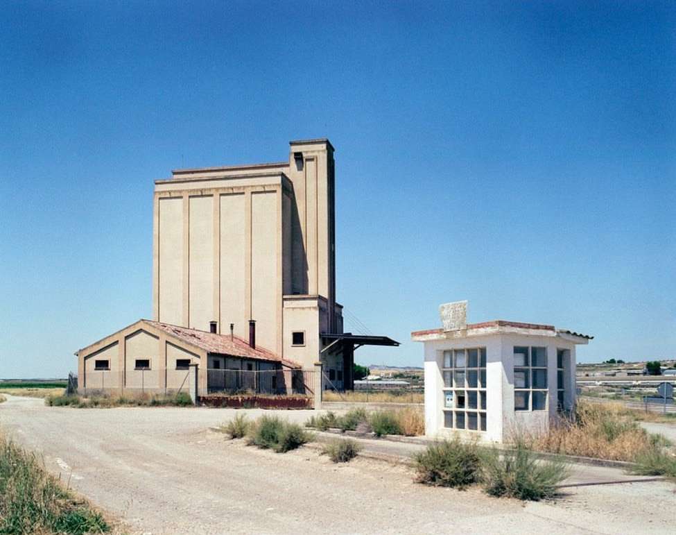 Daytime exterior shot of a tall beige grain elevator with a small white guardhouse, under a clear blue sky in a rural landscape.