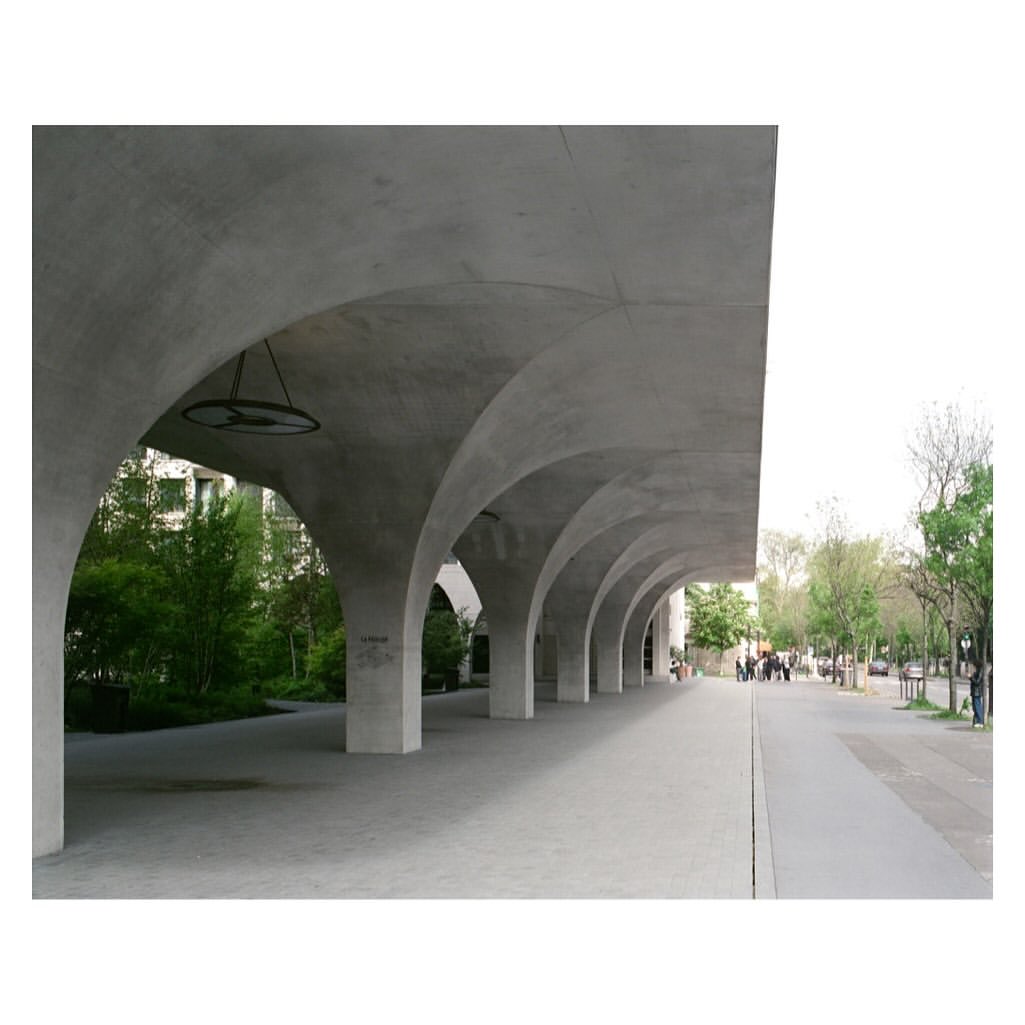 Concrete archway with supporting pillars, leading to a tree-lined walkway with distant figures and cars. Architectural design.