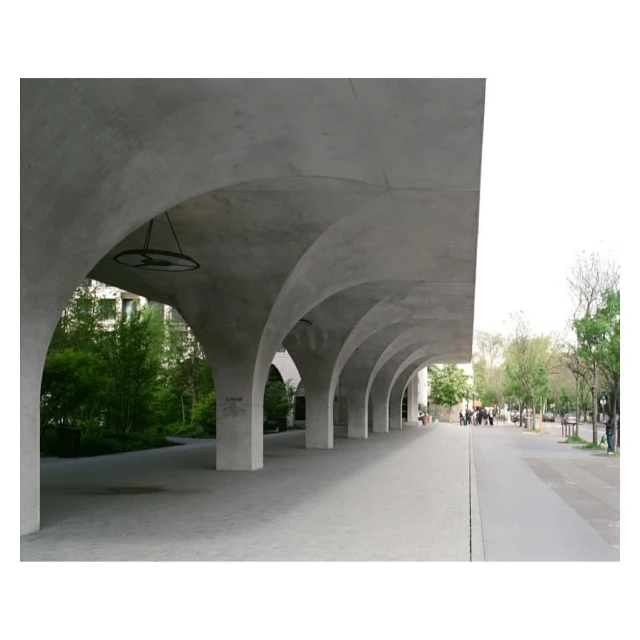 Concrete archway with supporting pillars, leading to a tree-lined walkway with distant figures and cars. Architectural design.
