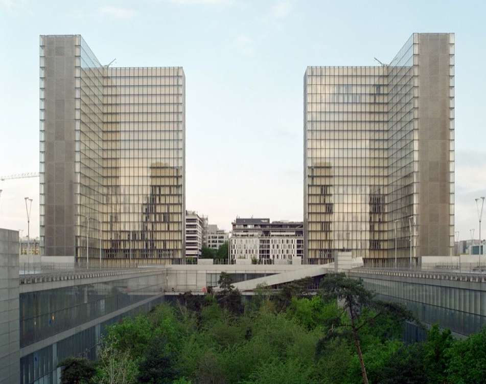 Exterior daytime view of the Bibliothèque nationale de France, two modern high-rise buildings with a bridge-like structure and green trees.