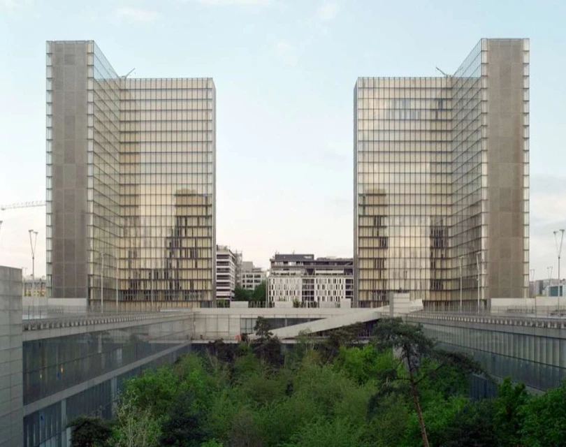 Exterior daytime view of the Bibliothèque nationale de France, two modern high-rise buildings with a bridge-like structure and green trees.