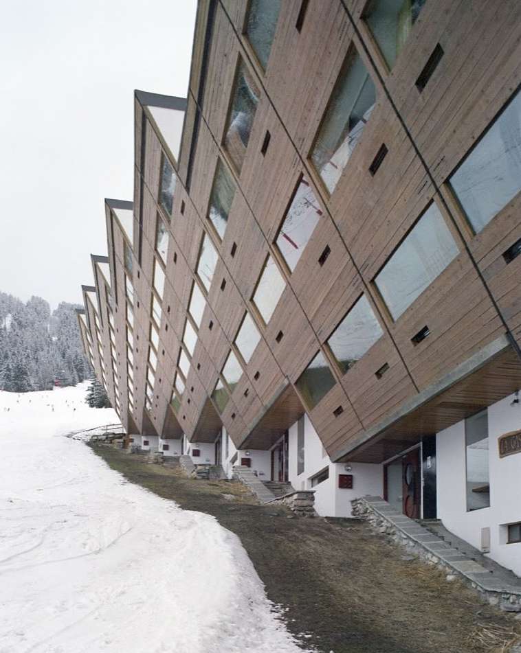 Exterior view of a modern geometric hotel with wood paneling and large windows, situated near a snow-covered ski slope and dense forest.