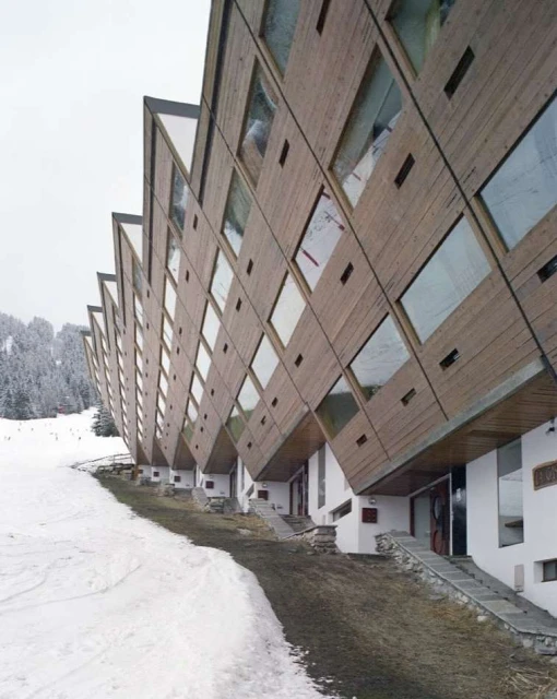 Exterior view of a modern geometric hotel with wood paneling and large windows, situated near a snow-covered ski slope and dense forest.