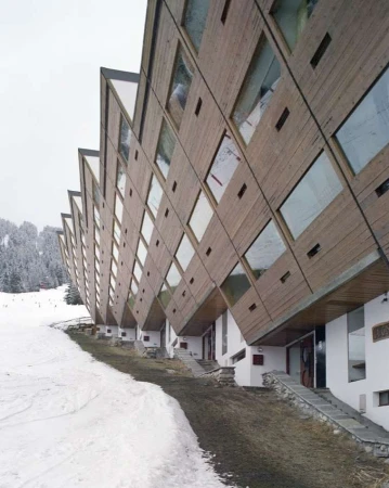 Exterior view of a modern geometric hotel with wood paneling and large windows, situated near a snow-covered ski slope and dense forest.