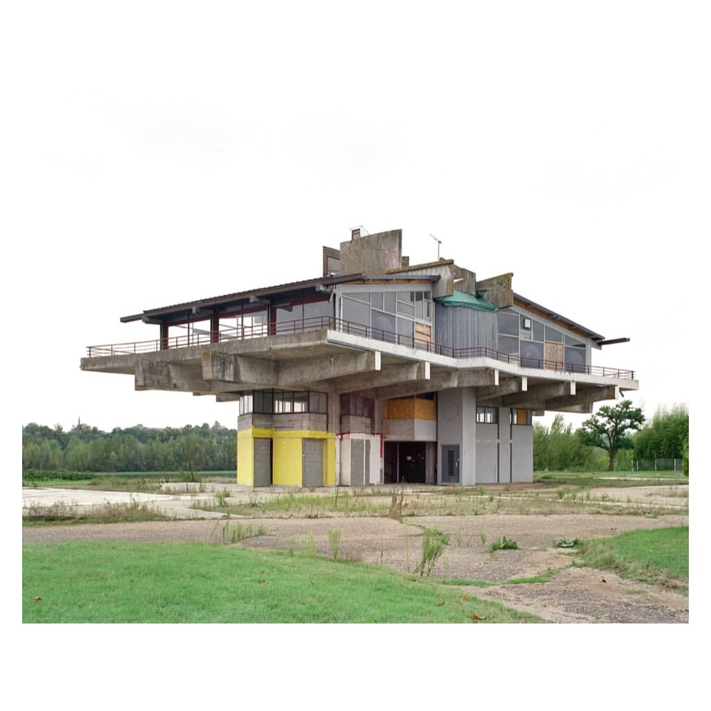 Exterior view of a unique concrete architecture with large overhanging balcony surrounded by grassy fields and a grey sky.