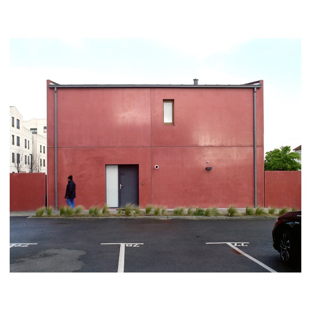 Modern red building with grey door and small window, a man walking in front, and parking spaces in the foreground.