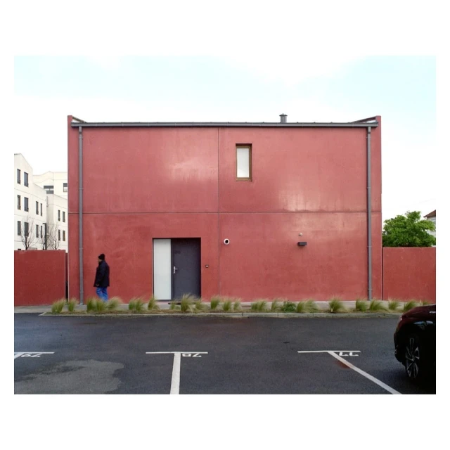 Modern red building with grey door and small window, a man walking in front, and parking spaces in the foreground.