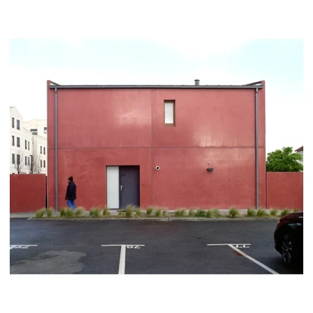 Modern red building with grey door and small window, a man walking in front, and parking spaces in the foreground.