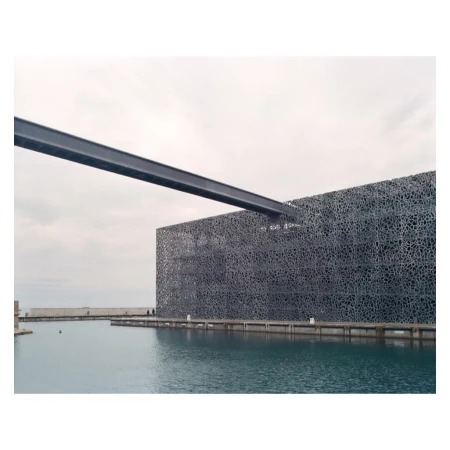 Exterior view of the MuCEM building in Marseille, France, featuring its unique textured facade, connected steel beam, and surrounding water.