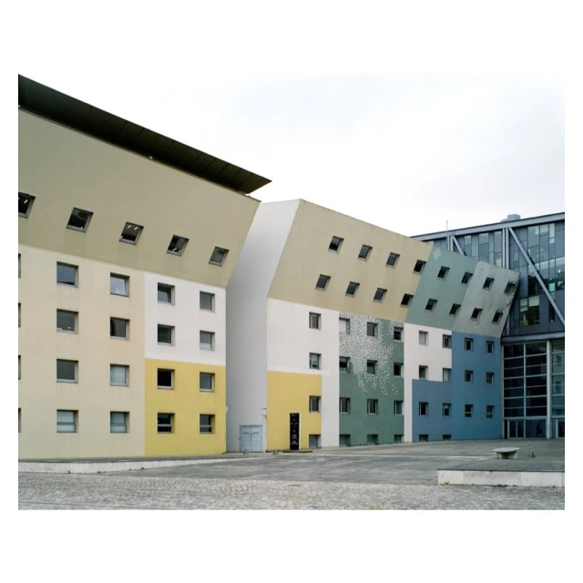 Exterior of a modern, colorful building featuring an irregular design with many windows, set against an overcast sky and paved courtyard.