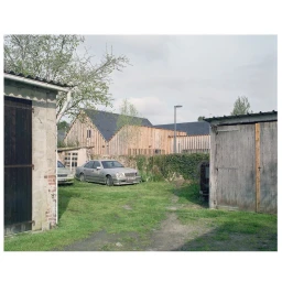 An outdoor courtyard view with weathered wooden outbuildings, parked sedans, a stone wall, and modern vertical wood clad buildings in Paris