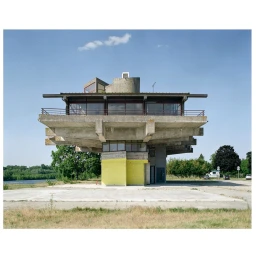 Brutalist concrete building with cantilevered floors and a yellow base under a clear blue sky, photographed by Fred Guillaud.