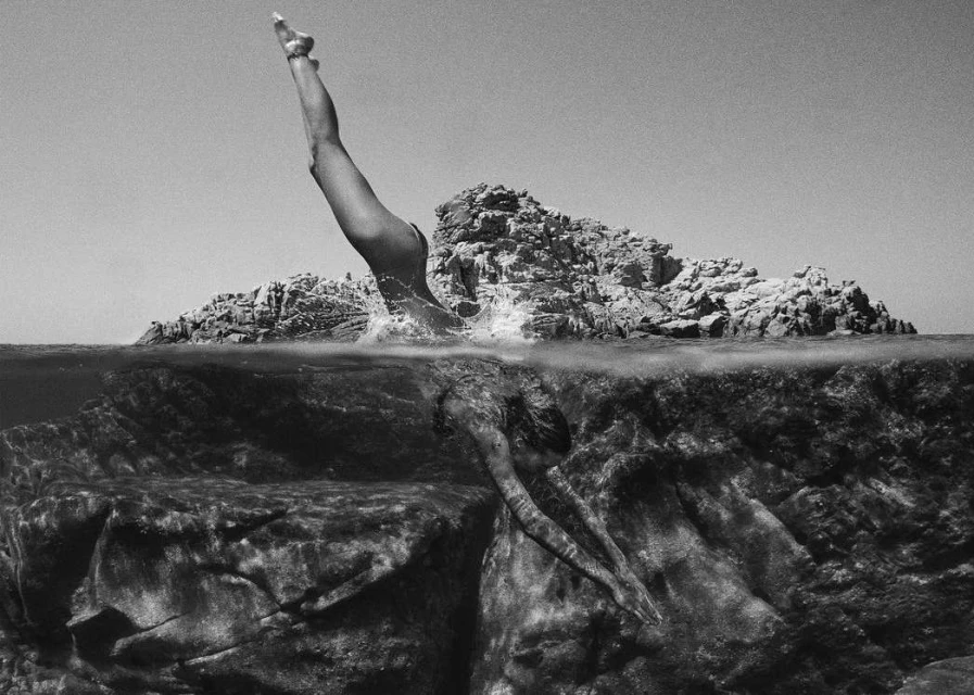 Black and white underwater view of a woman diving headfirst into the ocean, surrounded by rock formations and clear sky above.