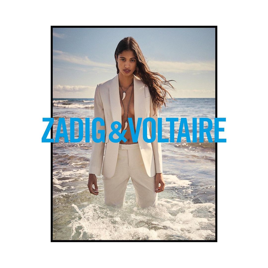 A fashion model stands in ocean water wearing a white pantsuit, featuring a white sandy beach, ocean, and sky in the background.