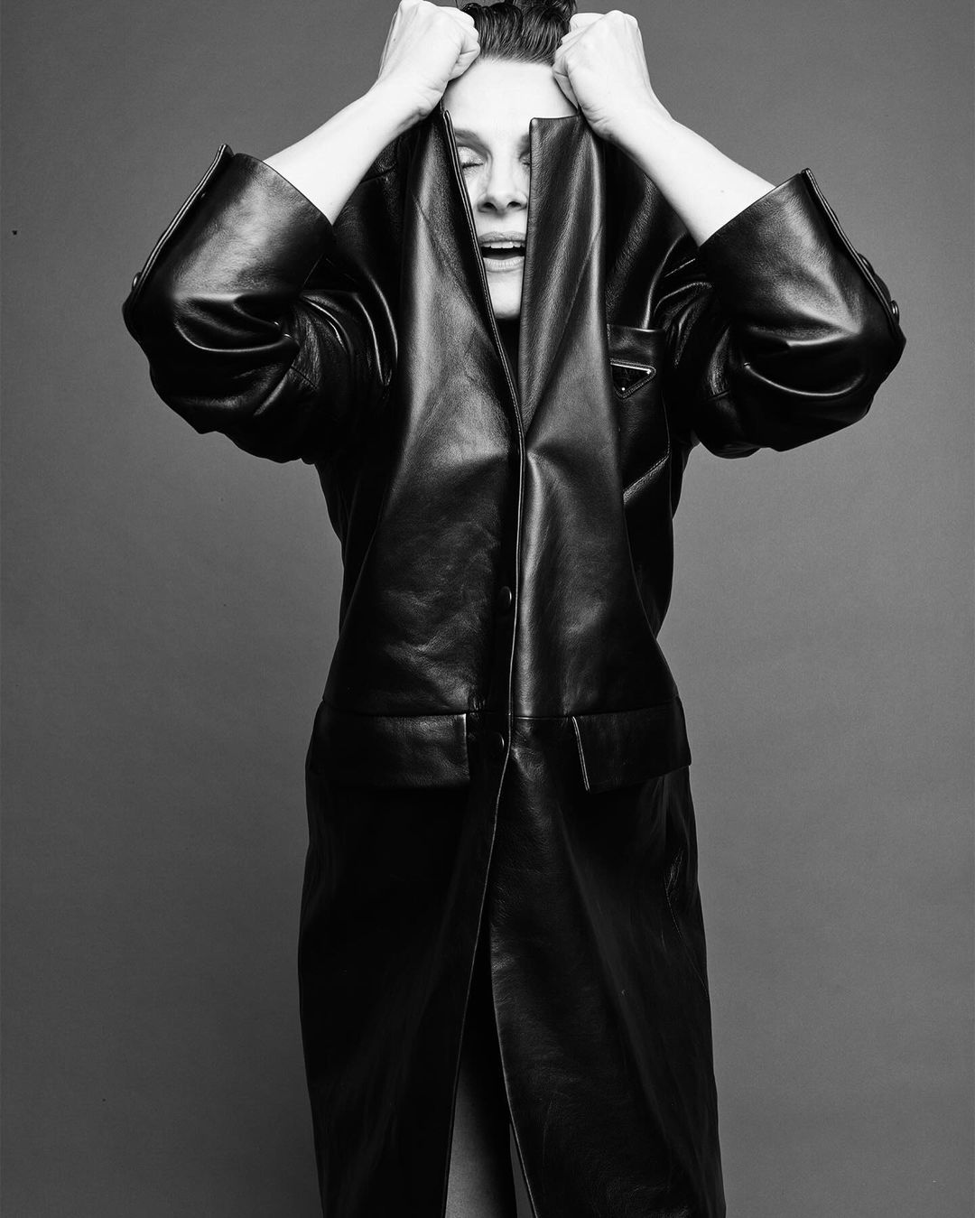 Monochrome fashion portrait of a woman in a black leather coat with her hands on her head against a gray backdrop in a photo studio.