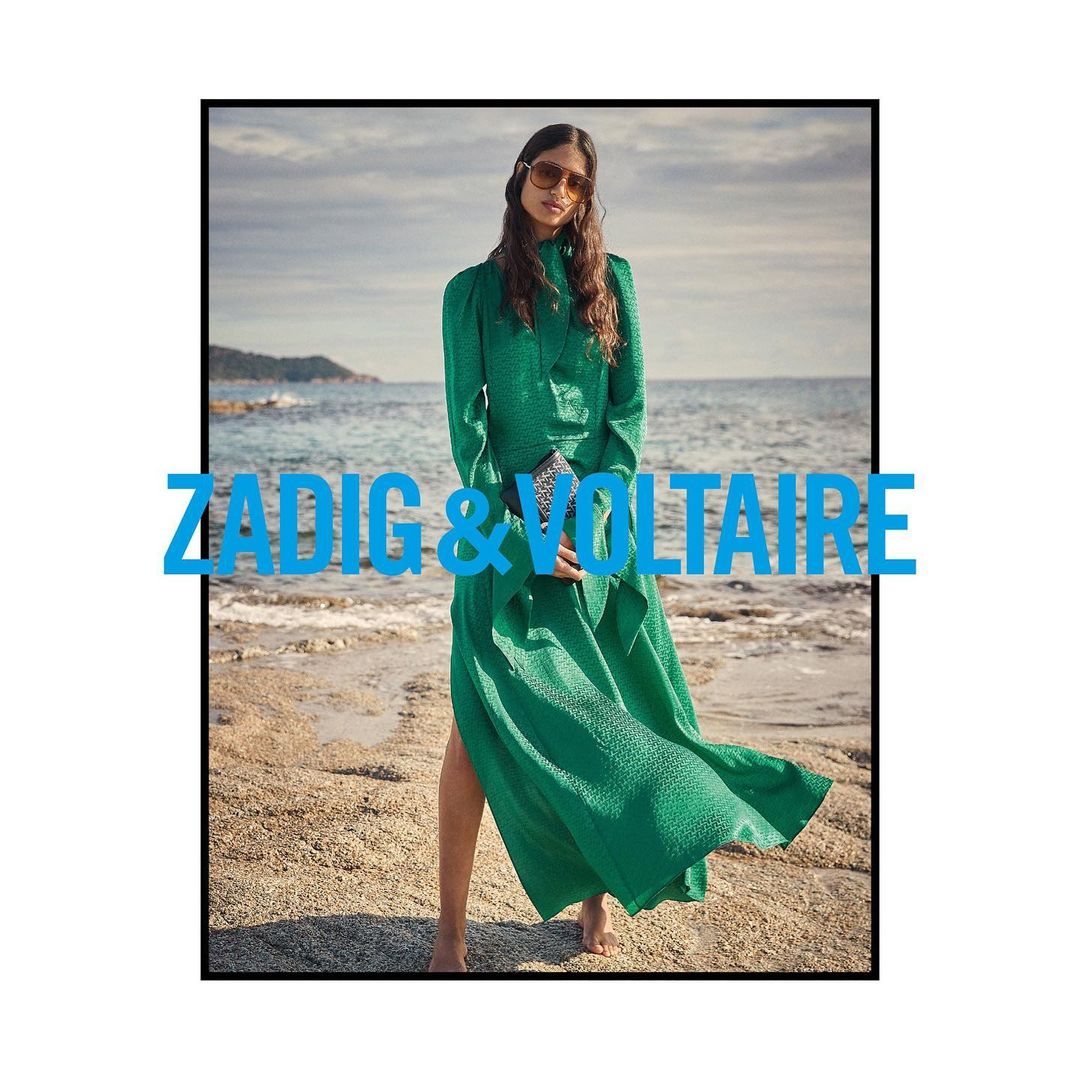 A fair-skinned adult woman wears a long-sleeve green dress and sunglasses, standing barefoot on a sandy beach next to the ocean.
