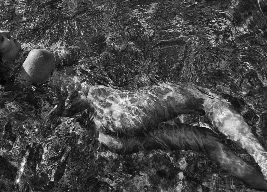 Black-and-white image of an adult woman lying in water with ripples and refractions visible on the surface and her body under the water.