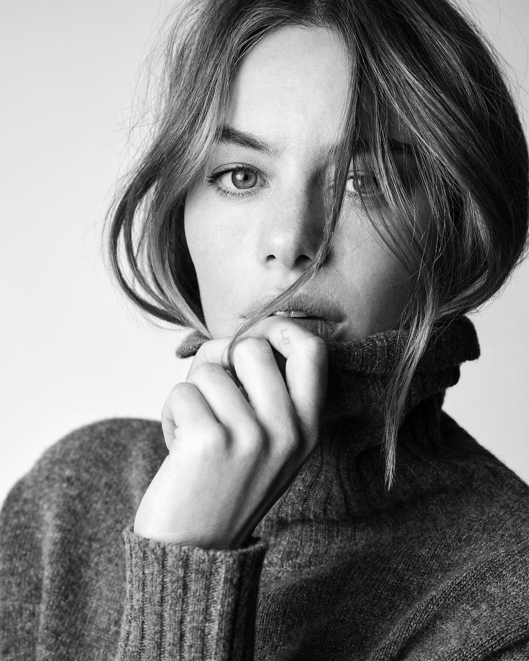 Black-and-white portrait of a woman with shoulder-length hair wearing a large turtleneck sweater, touching her face in a studio setting.