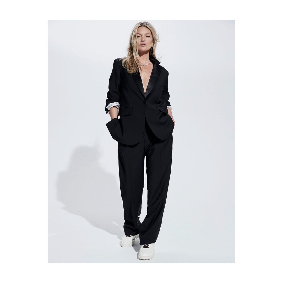 A blonde woman in a black blazer and pantsuit, hands in pockets, poses confidently against a white background in a fashion editorial.