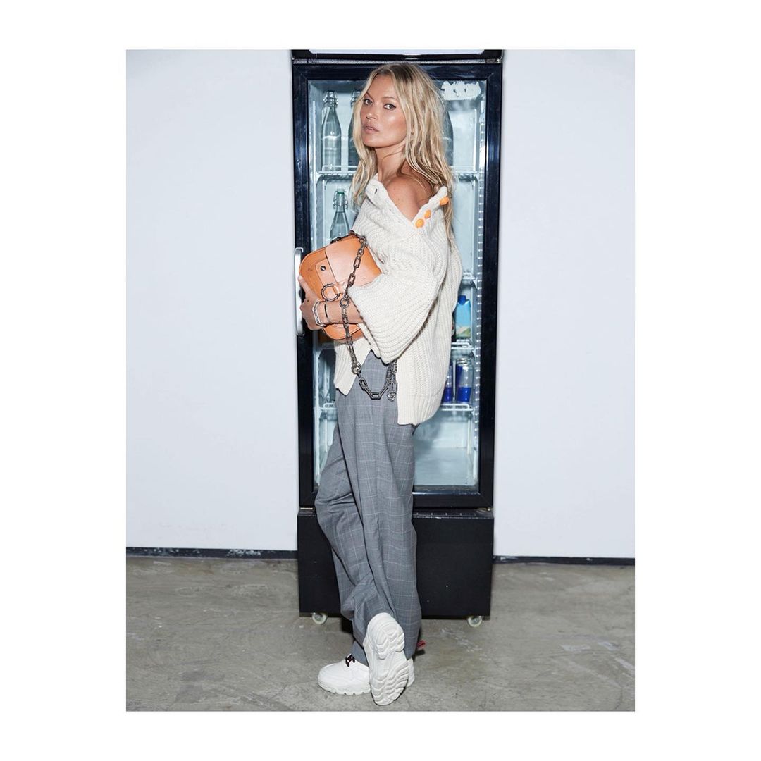 A blonde woman poses in front of a cooler holding a purse, wearing a cream sweater, grey pants, and white sneakers in a casual indoor shot.
