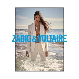 Woman wearing a cream suit standing waist-deep in ocean surf with blue text overlay reading ZADIG and VOLTAIRE