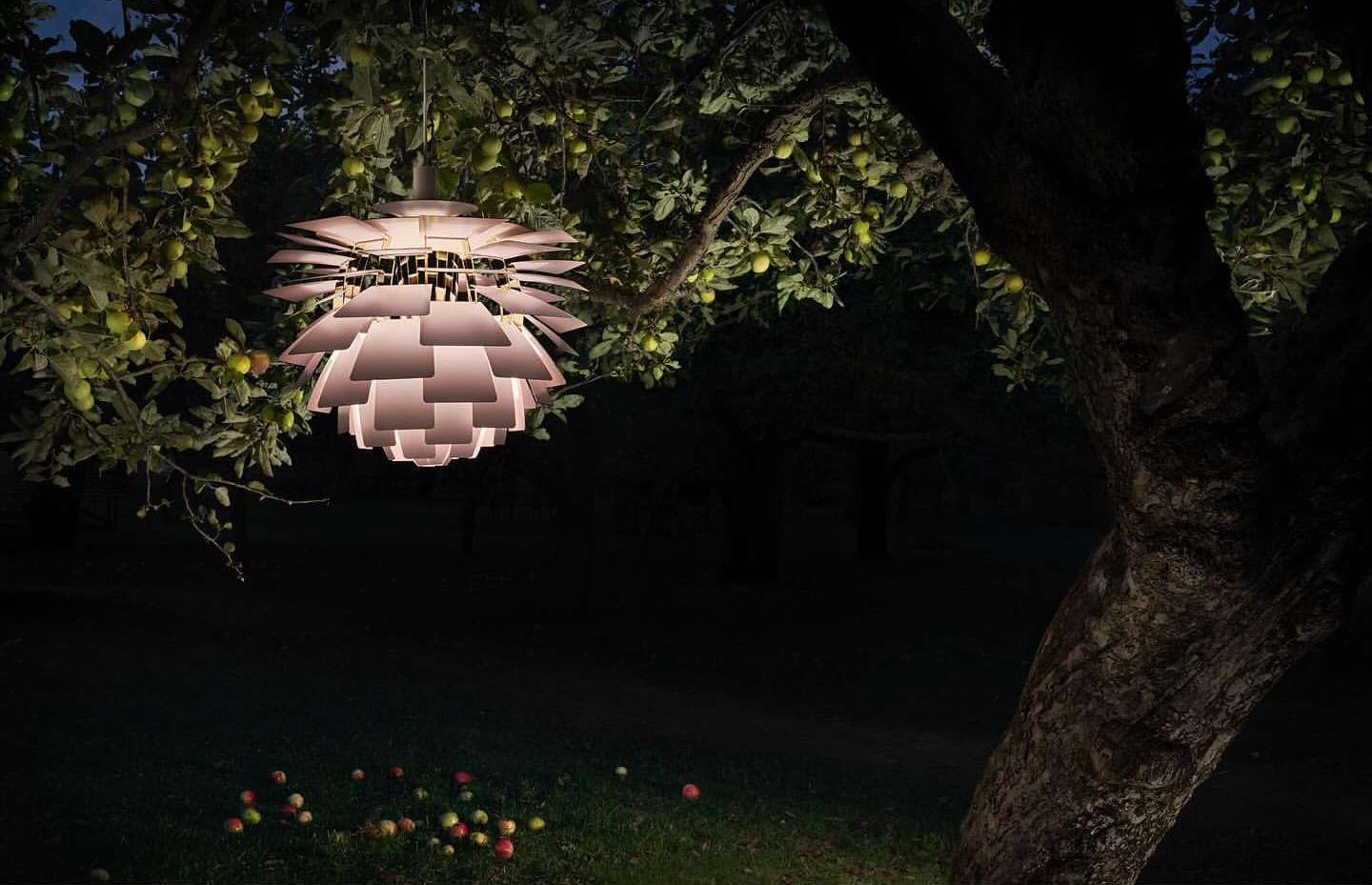 Illuminated pendant lamp hanging from a fruit tree at night, casting light on the apples scattered on the lawn below in a dark garden.