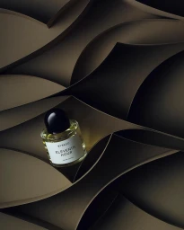 A clear glass perfume bottle with a black cap resting on layered brown curved paper shapes in moody lighting