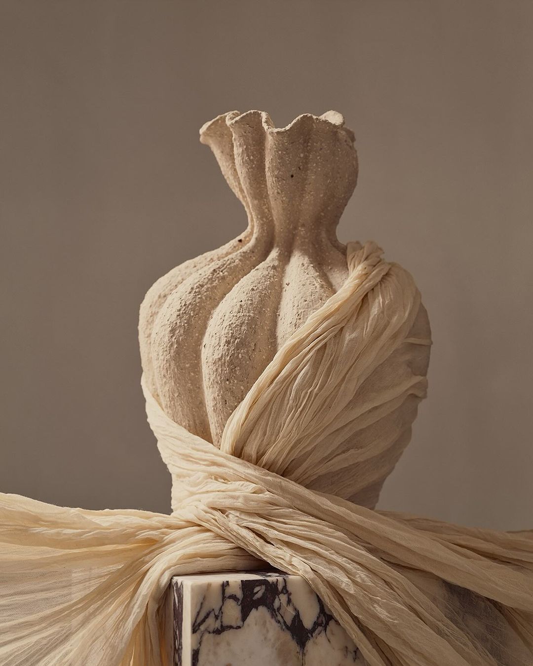 Elegant still life: Textured vase on a marble cube, draped with soft fabric, bathed in warm, inviting light against a neutral backdrop.