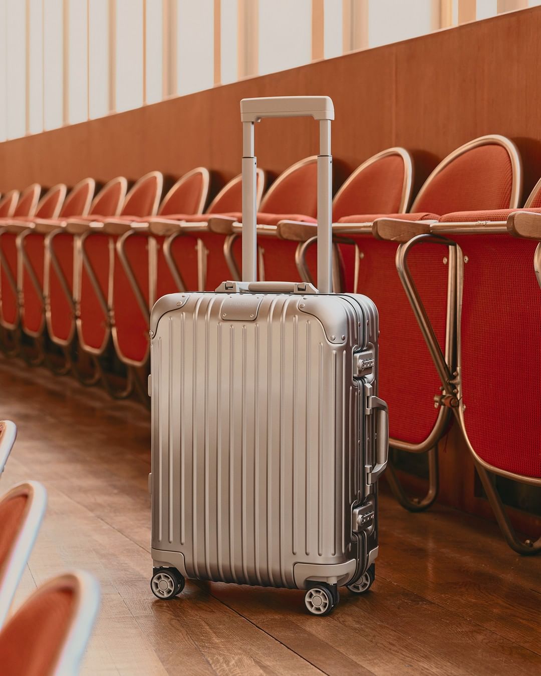 Silver rolling suitcase in an indoor space with a wooden floor and red cushioned seating in the background; commercial travel.