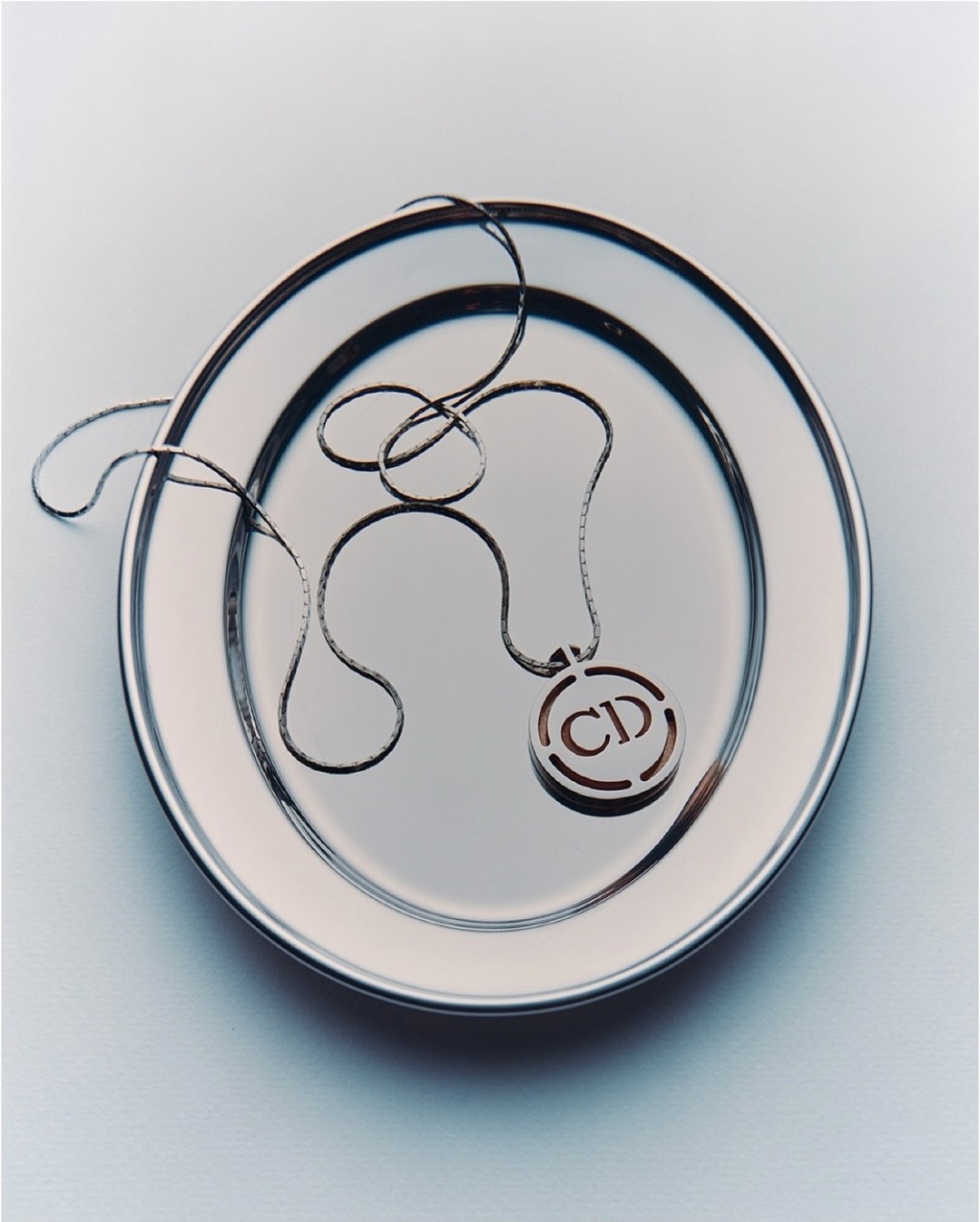 Silver Christian Dior logo pendant necklace on a shiny silver plate. Elegant studio product shot. Luxury fashion accessory.