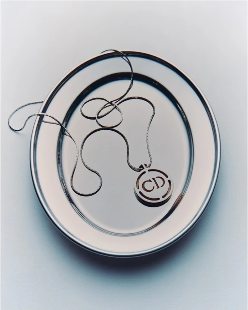 Silver Christian Dior logo pendant necklace on a shiny silver plate. Elegant studio product shot. Luxury fashion accessory.