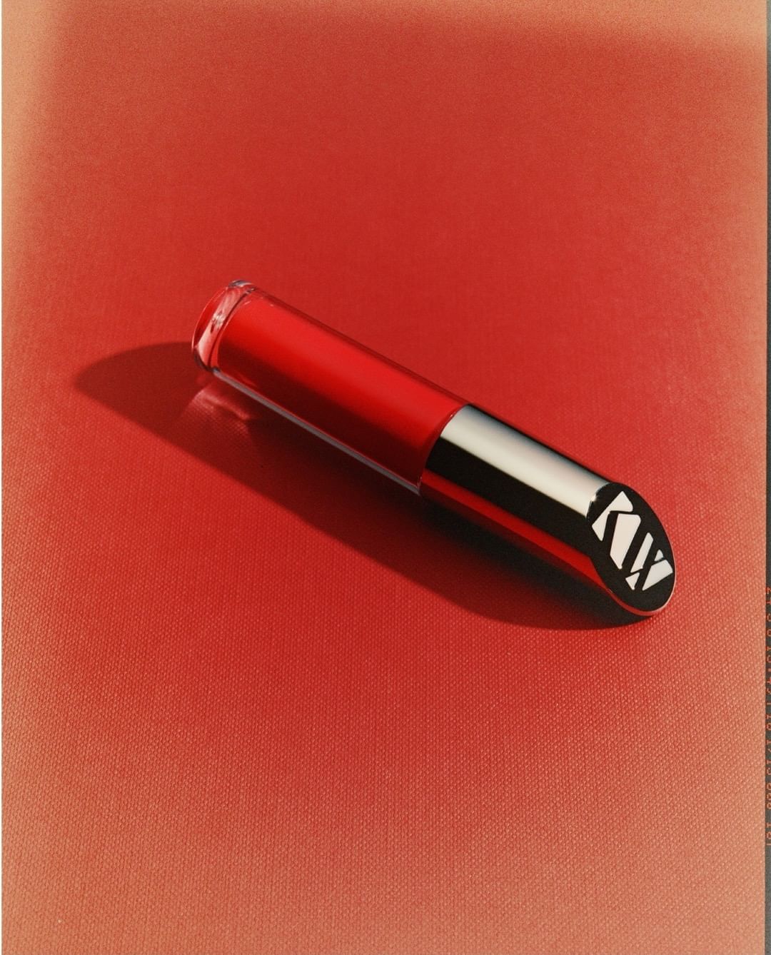 Red lip gloss tube with silver accents and patterned cap on a gradient red background, product shot for beauty and fashion.