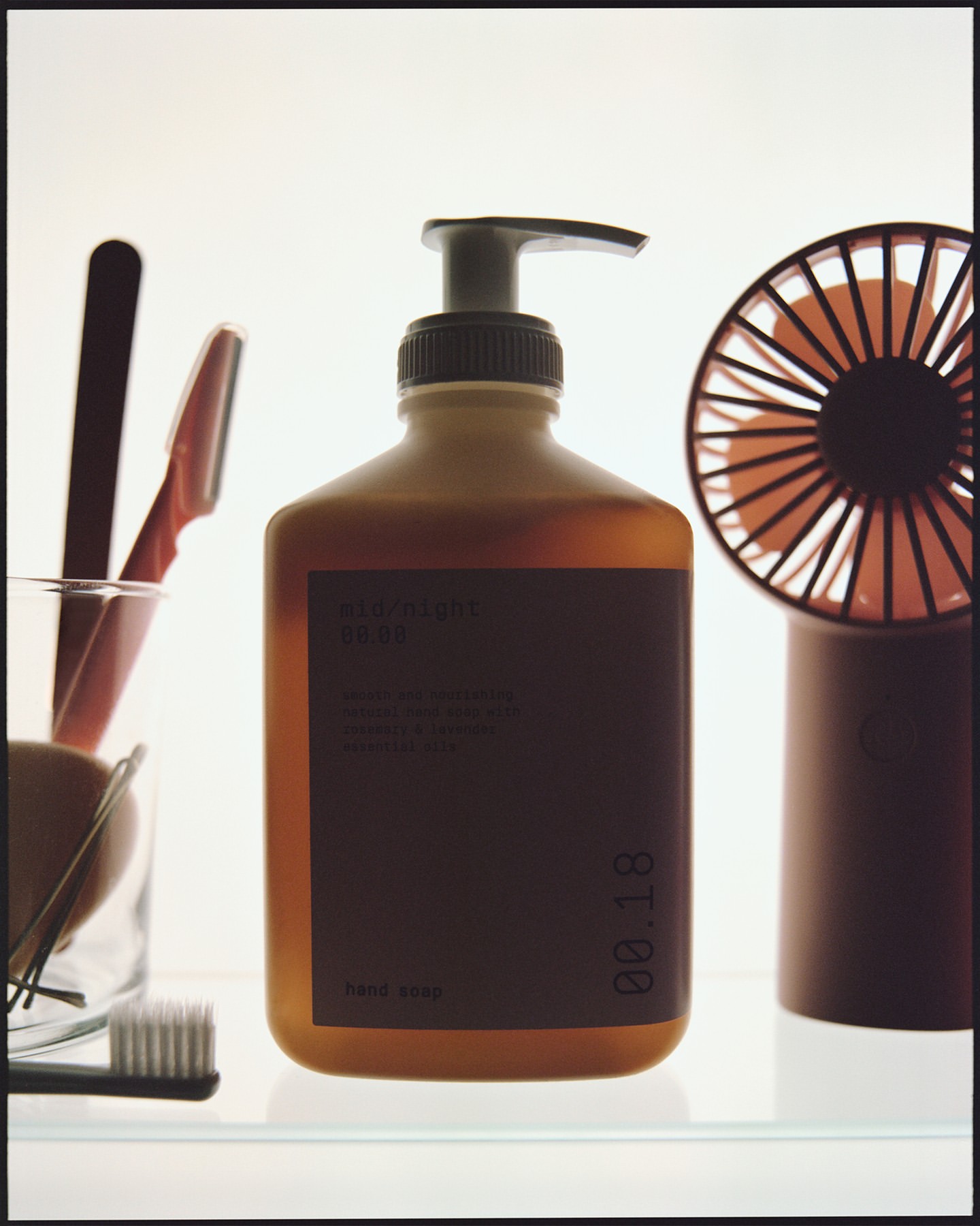 Close-up of an amber glass soap dispenser with a black label alongside a small fan and toiletries against a bright white background.