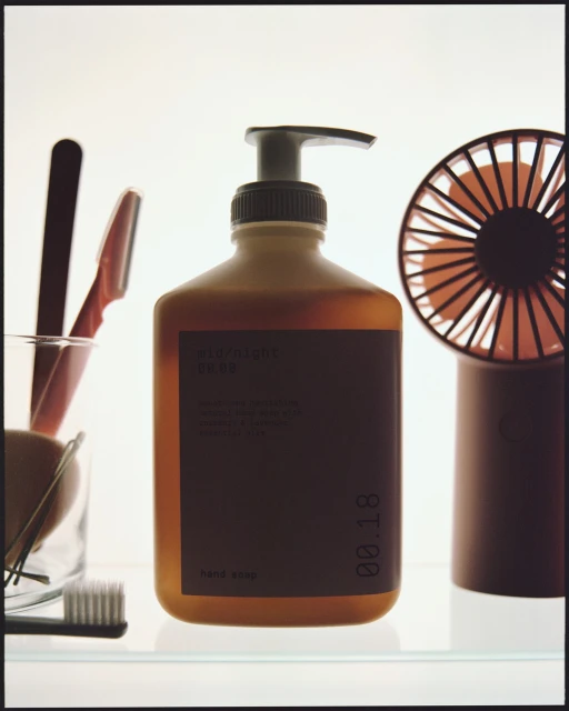 Close-up of an amber glass soap dispenser with a black label alongside a small fan and toiletries against a bright white background.