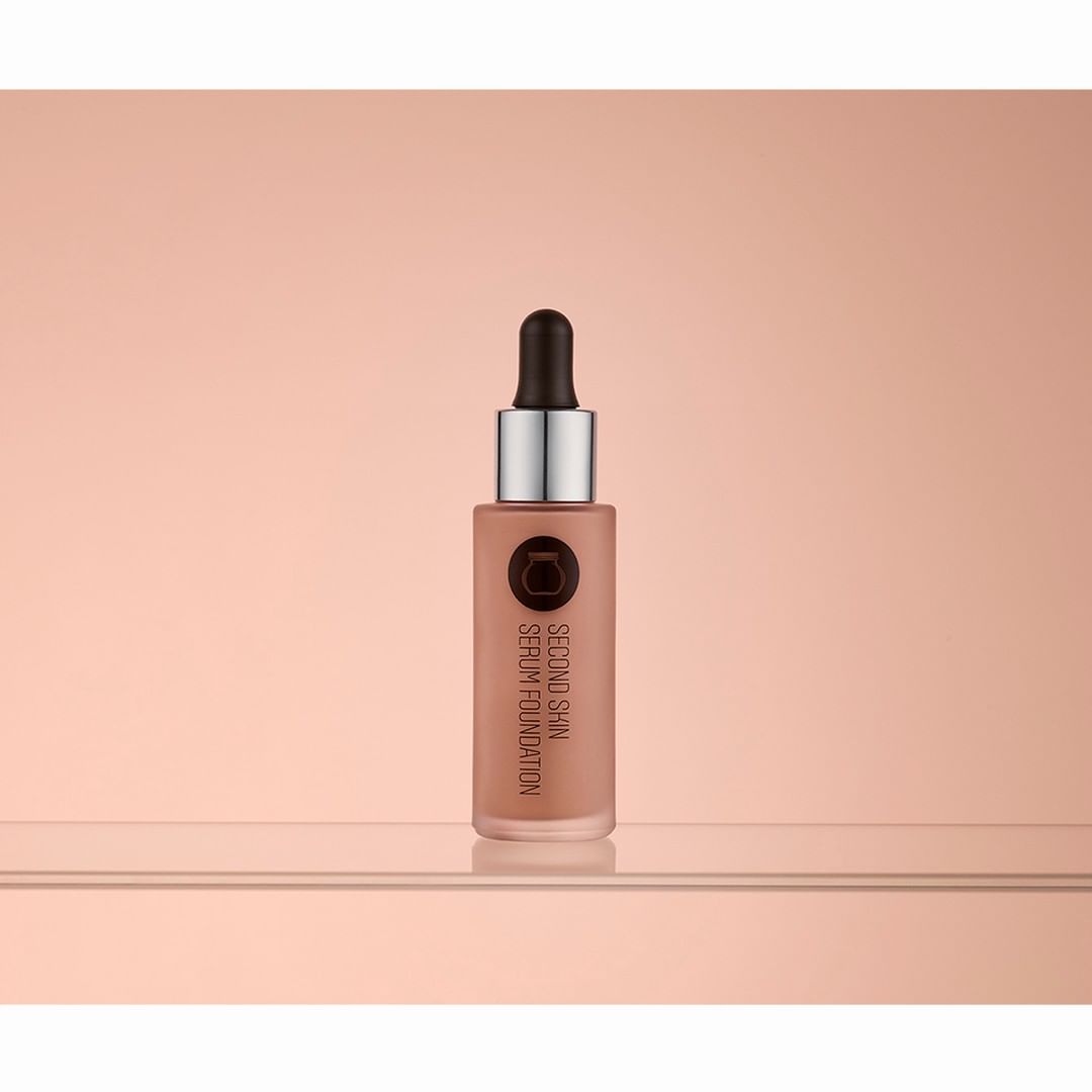 Studio shot of a bottle of 'Second Skin' serum foundation on a reflective surface with a diffused peach-toned background.