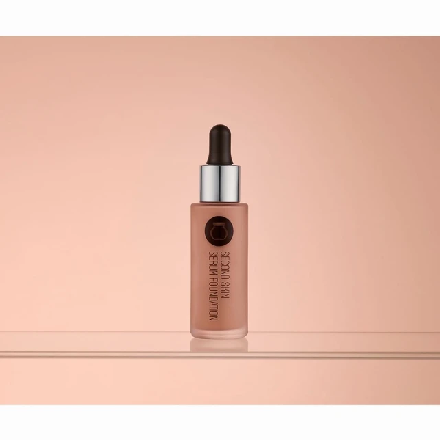 Studio shot of a bottle of 'Second Skin' serum foundation on a reflective surface with a diffused peach-toned background.