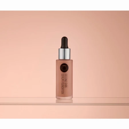 Studio shot of a bottle of 'Second Skin' serum foundation on a reflective surface with a diffused peach-toned background.