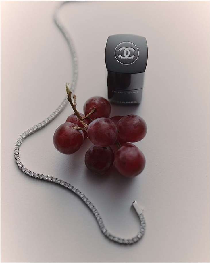Still life shot of black Chanel skincare container next to red grapes and a diamond necklace on a white surface in a studio setting.