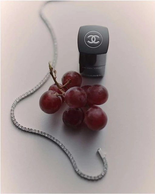 Still life shot of black Chanel skincare container next to red grapes and a diamond necklace on a white surface in a studio setting.