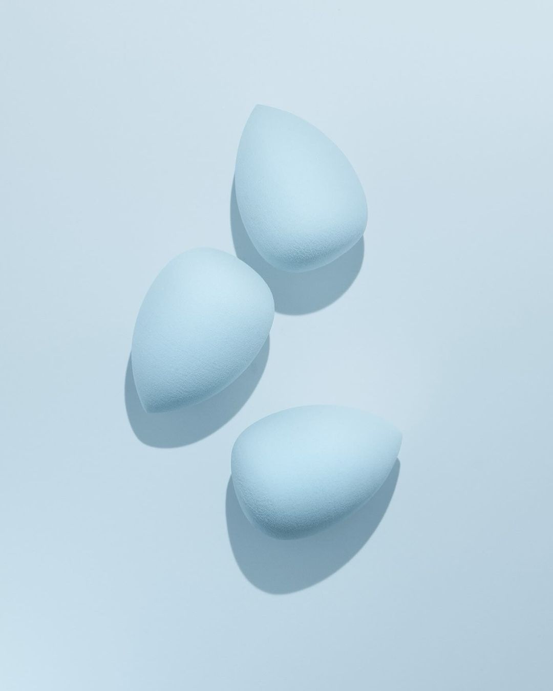 Three light blue makeup blending sponges arranged on a light blue surface, casting soft shadows in a beauty product still life.
