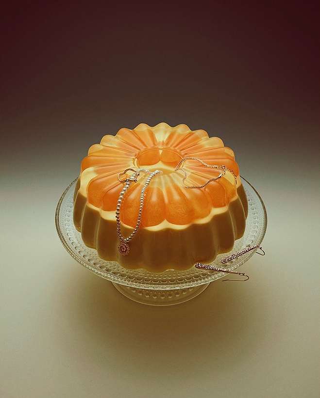 Still life photo of a two-tiered gelatin dessert on a stemmed glass plate with jewelry draped across the top tier, studio lighting.