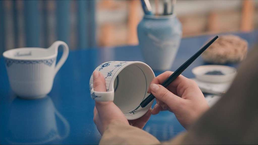 Close-up of artisan's hands painting blue floral pattern on white porcelain tea cup with paintbrush at blue tabletop indoors.
